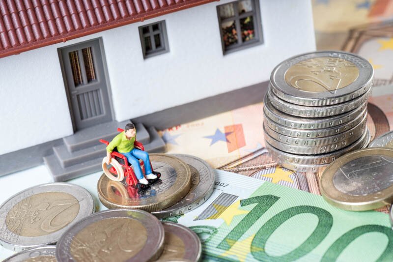 Rollstuhlfahrer vor Modellhaus auf Geldscheinen – symbolisiert behinderungsbedingte Ausgaben und Wohnkosten